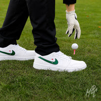 Golfer wearing white Air Jordan 1 Low Golf shoes with green golf-themed Swooshes, standing beside a golf ball on a pink tee.