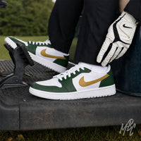 Golfer wearing white Air Jordan 1 Low Golf shoes with dark green overlays and gold Nike Swooshes, seated in a golf cart with a white-and-black golf glove.