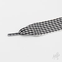 Close-up of a wide black-and-white gingham shoelace with frilled edges and a clear plastic aglet on a white background.