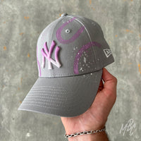 Gradient Swipe custom New Era New York Yankees cap featuring a grey crown, pink ombré embroidered NY logo, hand-painted purple gradient swipe graphics and white paint splatter detailing, held in hand.
