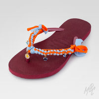 Angled view of a burgundy Havaianas flip-flop featuring orange and light blue braided straps, fabric bows and daisy, cross and evil-eye charms.