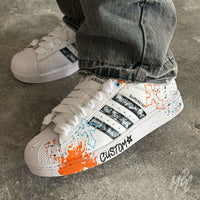 Modern Relic custom Adidas Superstar worn on foot, showcasing textured black paint stripes, orange and blue graffiti-inspired artwork, hand-painted paint splatters and white rope laces.
