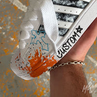 Close-up of the Modern Relic custom Adidas Superstar highlighting textured black paint stripes, orange paint splatters, blue graffiti details, hand-painted midsole artwork and white rope laces.