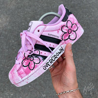 Hand holding Pink Overgrown custom Adidas Superstar featuring hand-painted pink flowers, textured paint finish and Overgrown branding.