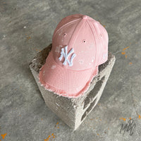 Pink Raw Edge custom New Era New York Yankees cap displayed on a concrete block, featuring a distressed frayed brim, frayed white NY logo, white paint splatter accents and a soft pink finish.