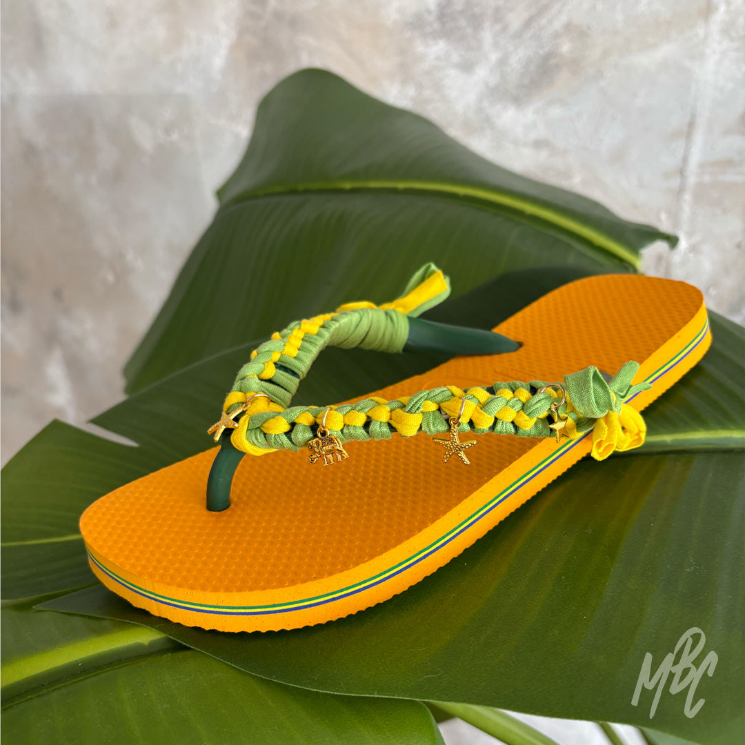 Sunwoven Carnival custom Havaianas featuring handcrafted yellow and green braided straps with gold starfish and turtle charms resting on banana tree leaf