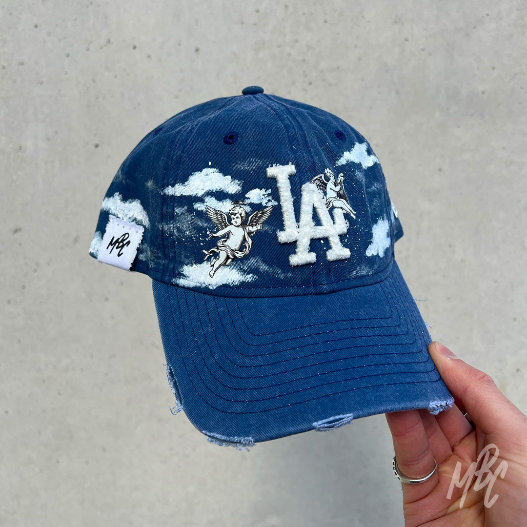Blue denim New Era LA custom cap with angel cherub illustrations and painted cloud design shown in hand