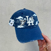 Blue denim New Era LA custom cap with angel cherub illustrations and painted cloud design shown in hand