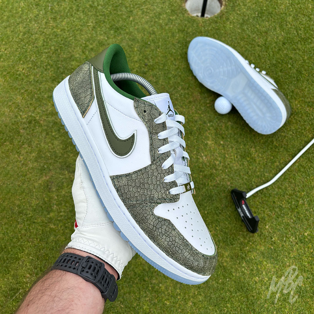 Emerald Reserve Jordan 1 Low Golf custom golf shoe held above a putting green with golf equipment in the background