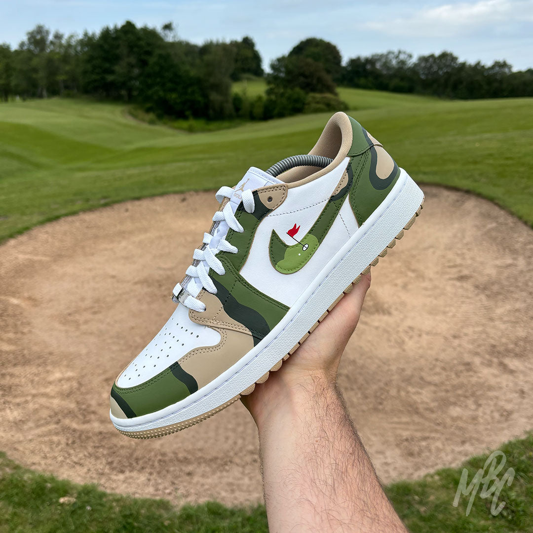 Hand holding a Fairway Camo Jordan 1 Low Golf custom golf trainer above a golf course bunker