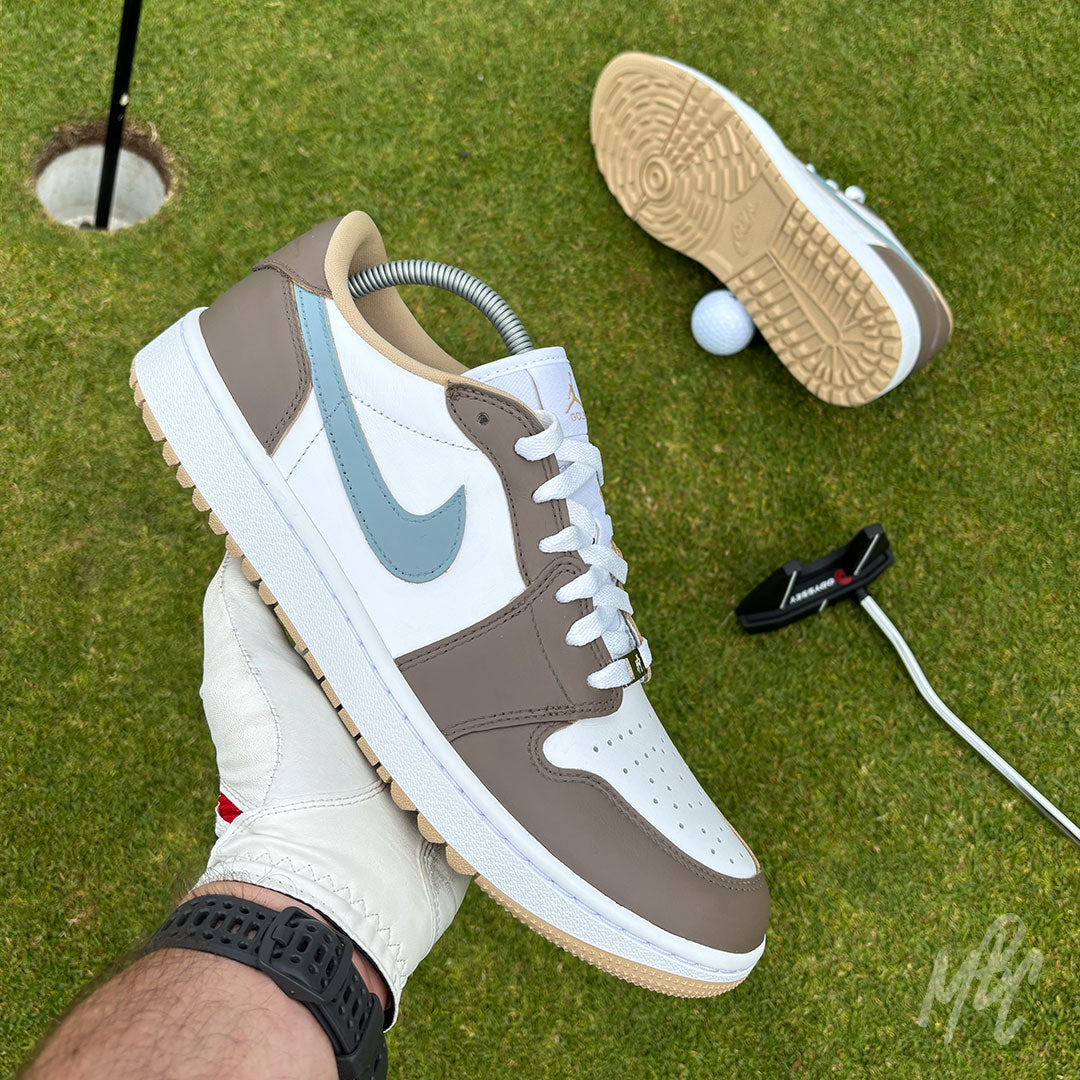 Fairway Fog Jordan 1 Low Golf custom golf shoe held above a putting green beside a golf hole, ball and putter