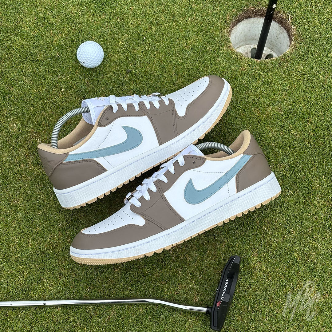 Pair of Fairway Fog Jordan 1 Low Golf custom golf trainers displayed on a putting green with golf ball and putter