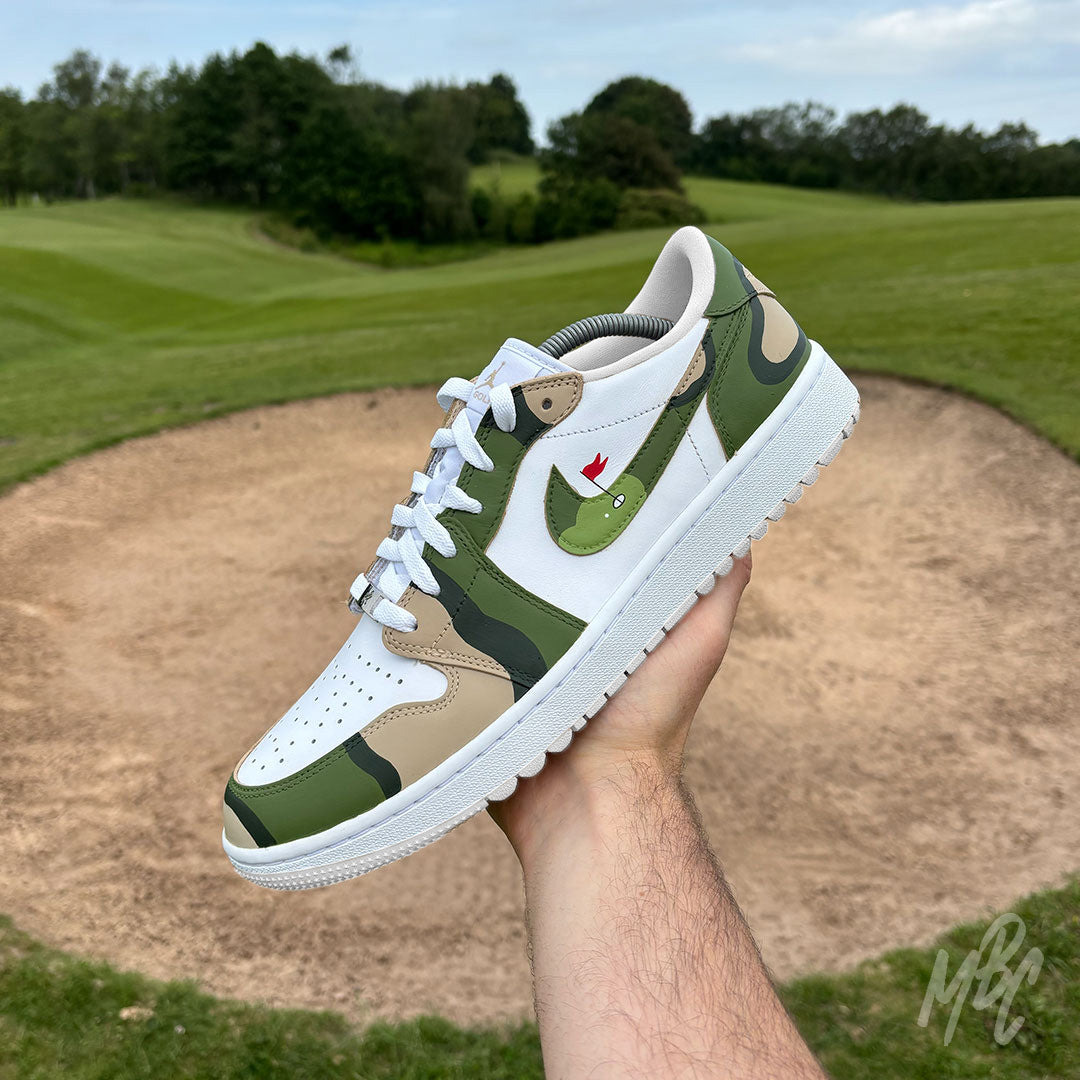 Hand holding a Fairway Camo Jordan 1 Low Golf custom golf trainer above a golf course bunker