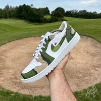 Hand holding a Fairway Camo Jordan 1 Low Golf custom golf trainer above a golf course bunker