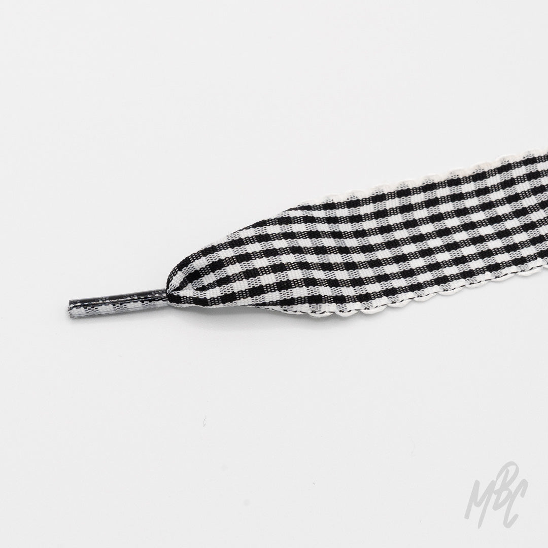 Close-up of a wide black-and-white gingham shoelace with frilled edges and a clear plastic aglet on a white background.