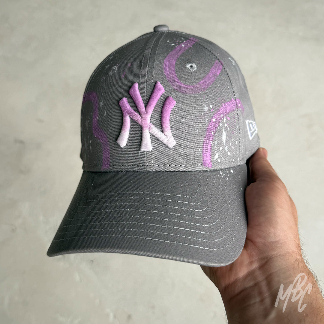 Front view of the Gradient Swipe custom New Era New York Yankees cap with a grey crown, pink gradient embroidered NY logo, purple hand-painted swipe graphics and white paint splatter finish.