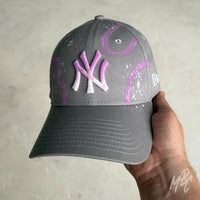Front view of the Gradient Swipe custom New Era New York Yankees cap with a grey crown, pink gradient embroidered NY logo, purple hand-painted swipe graphics and white paint splatter finish.