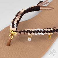 Close-up of the neutral braided straps on a brown Havaianas flip-flop, decorated with gold bow, pearl and letter charms.