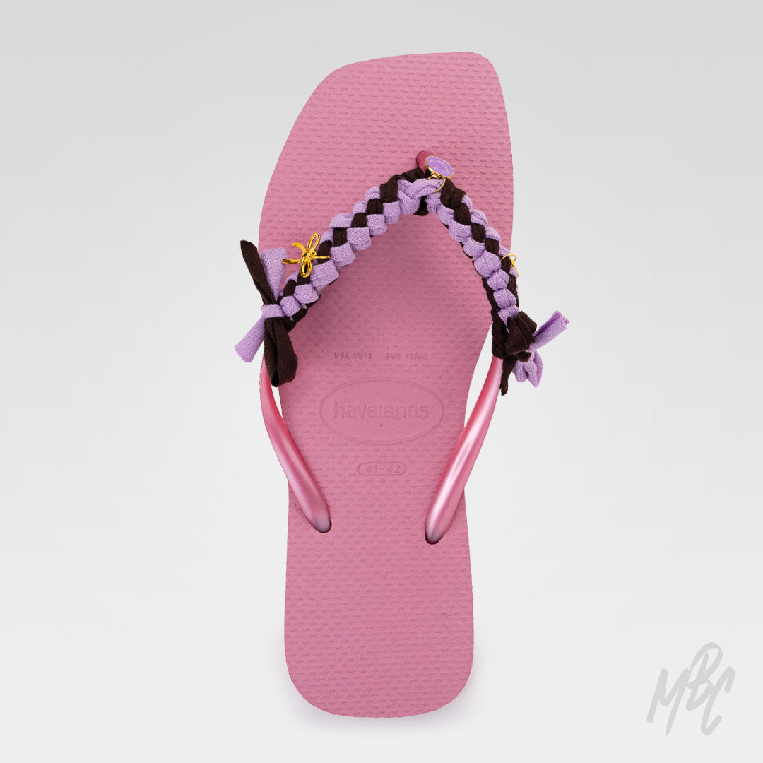 Top view of a pink Havaianas flip-flop featuring lilac and dark brown braided straps with fabric bows and gold charms.