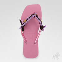 Top view of a pink Havaianas flip-flop featuring lilac and dark brown braided straps with fabric bows and gold charms.