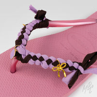 Close-up of a pink Havaianas flip-flop customised with lilac and dark brown braided straps, fabric bows and gold smiley face and bow charms.