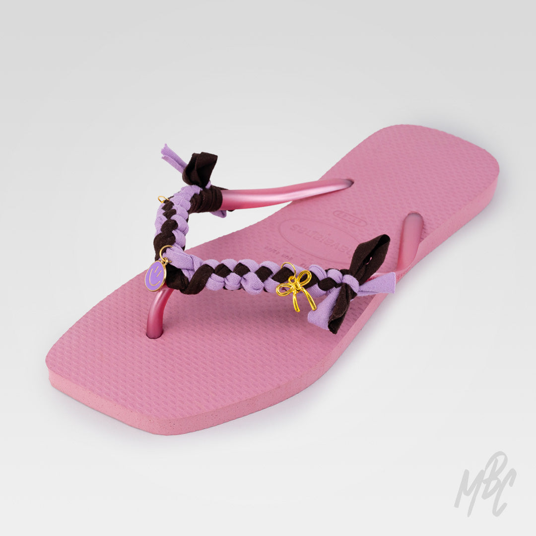 Angled view of a pink Havaianas flip-flop customised with lilac and dark brown braided straps, fabric bows and gold smiley face and bow charms.