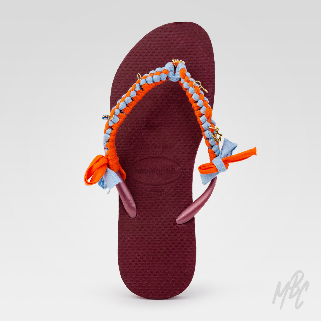 Top view of a burgundy Havaianas flip-flop customised with orange and light blue braided straps, fabric bows and gold charms.