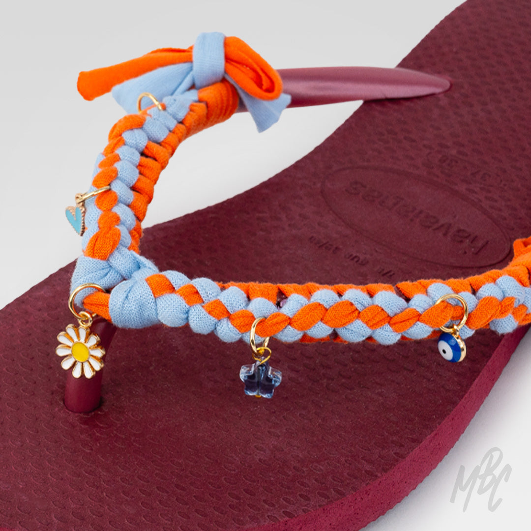 Close-up of the orange and light blue braided straps on a burgundy Havaianas flip-flop, decorated with daisy, blue cross and evil-eye charms.