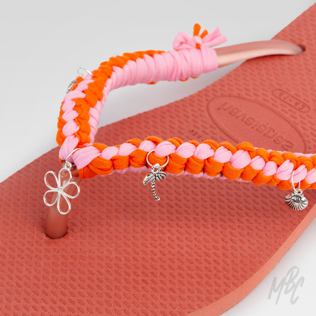Close-up of the pink and orange braided straps on a peach Havaianas flip-flop, decorated with silver flower, palm tree and shell charms.