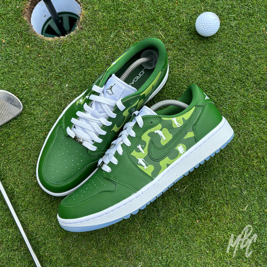 Pair of Lines of Play custom Jordan 1 Low Golf shoes with hand-painted golf-inspired camouflage artwork and golfer silhouettes displayed on a golf green beside a golf ball and club.