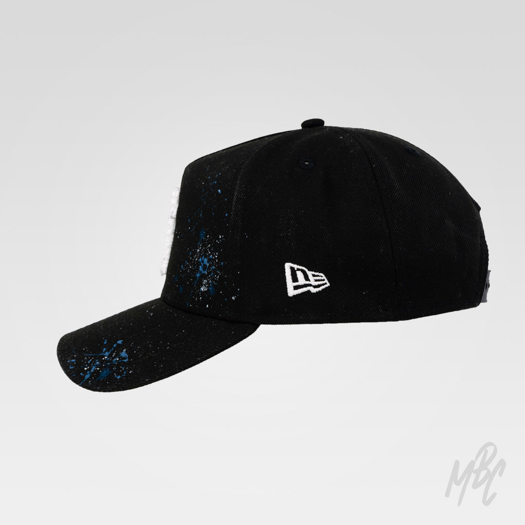 Side profile of the Midnight Canvas custom New Era cap featuring distressed white embroidered LA logo, hand-painted blue paint splatters and a black curved-brim design.