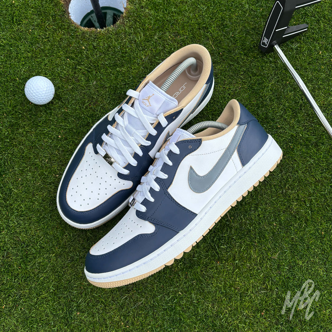 Pair of Midnight Tour custom Jordan 1 Low Golf shoes with premium white and grey leather with a navy blue hand painted colourway, photographed on a putting green beside a golf ball and putter.