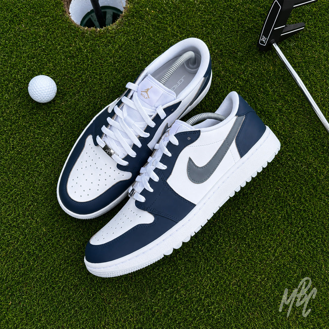 Pair of Midnight Tour custom Jordan 1 Low Golf shoes with premium white and grey leather with a navy blue hand painted colourway, photographed on a putting green beside a golf ball and putter.