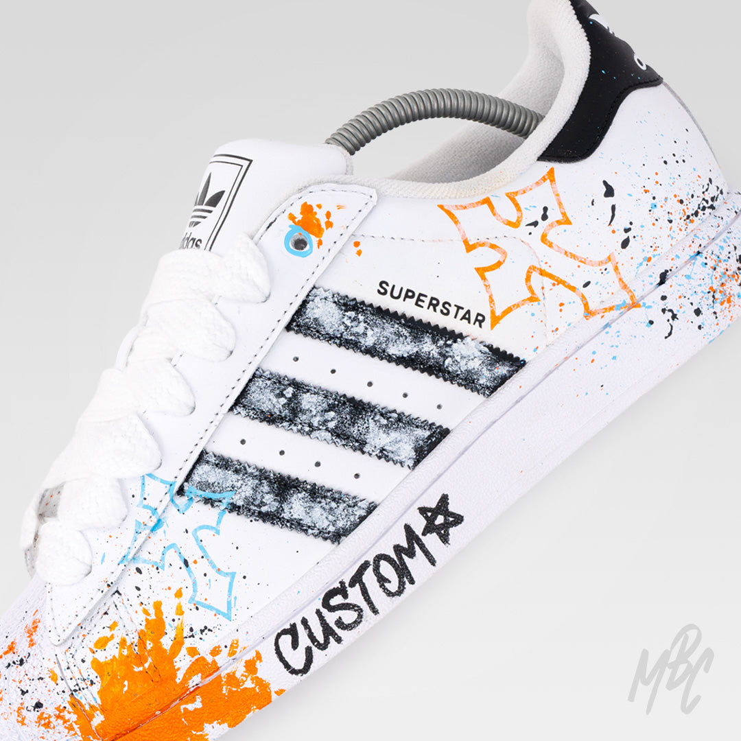 Close-up of Modern Relic custom Adidas Superstar featuring textured black painted stripes, hand-painted orange and blue graphics, paint splatters and graffiti-inspired artwork.