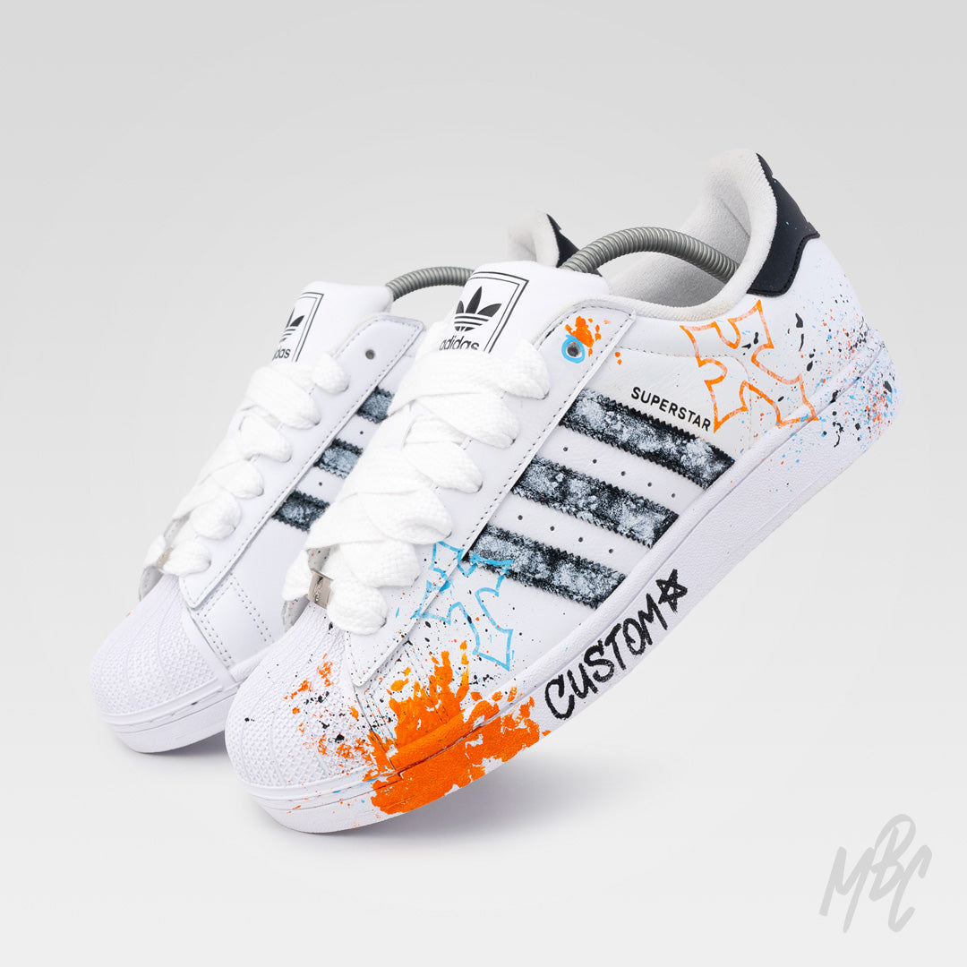 Pair of Modern Relic custom Adidas Superstar trainers featuring textured black painted stripes, hand-painted graffiti artwork and orange and blue paint splatters.