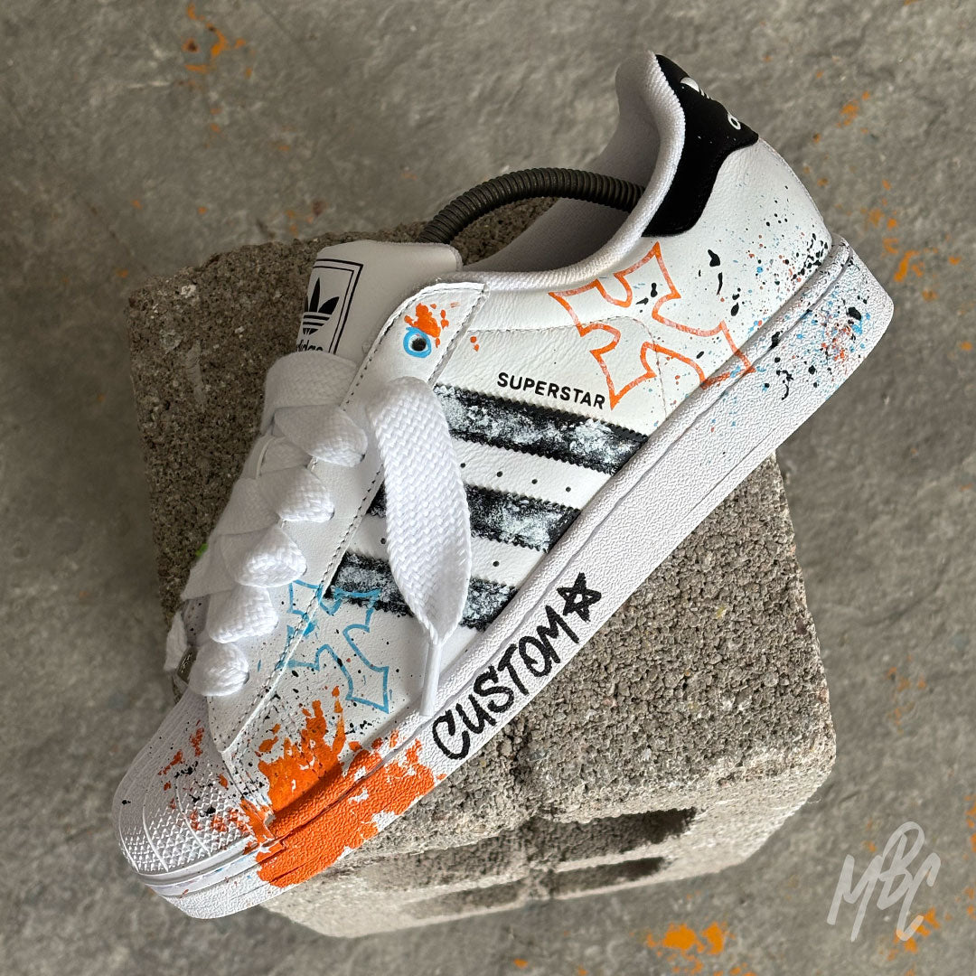 Modern Relic custom Adidas Superstar trainers featuring textured black paint stripes, hand-painted orange and blue graffiti details, paint splatters, white rope laces and hand-drawn artwork.