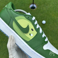 Pin Position custom Jordan 1 Low Golf shoes featuring hand-painted green fairway artwork on white leather, photographed on a putting green with a golf ball and hole.