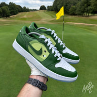 Pin Position custom Jordan 1 Low Golf shoes held above a golf green, featuring hand-painted fairway-inspired artwork on premium white and green leather.