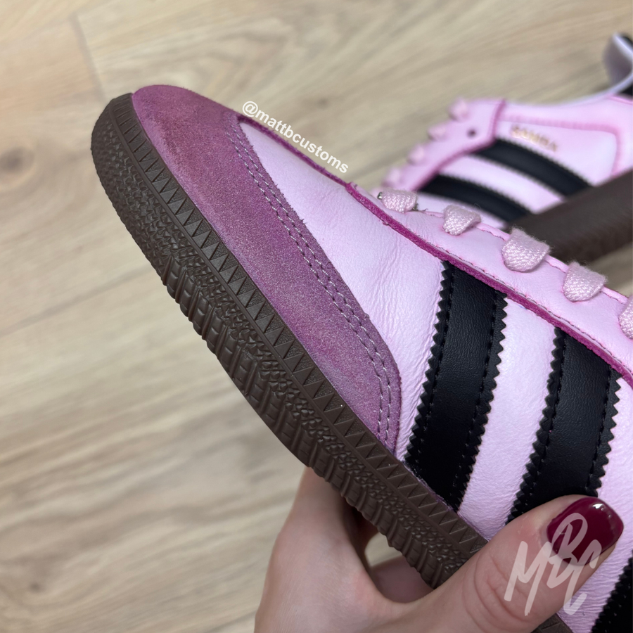 Close-up of a pink and black striped sneaker with brown sole held by a hand on a wooden floor.