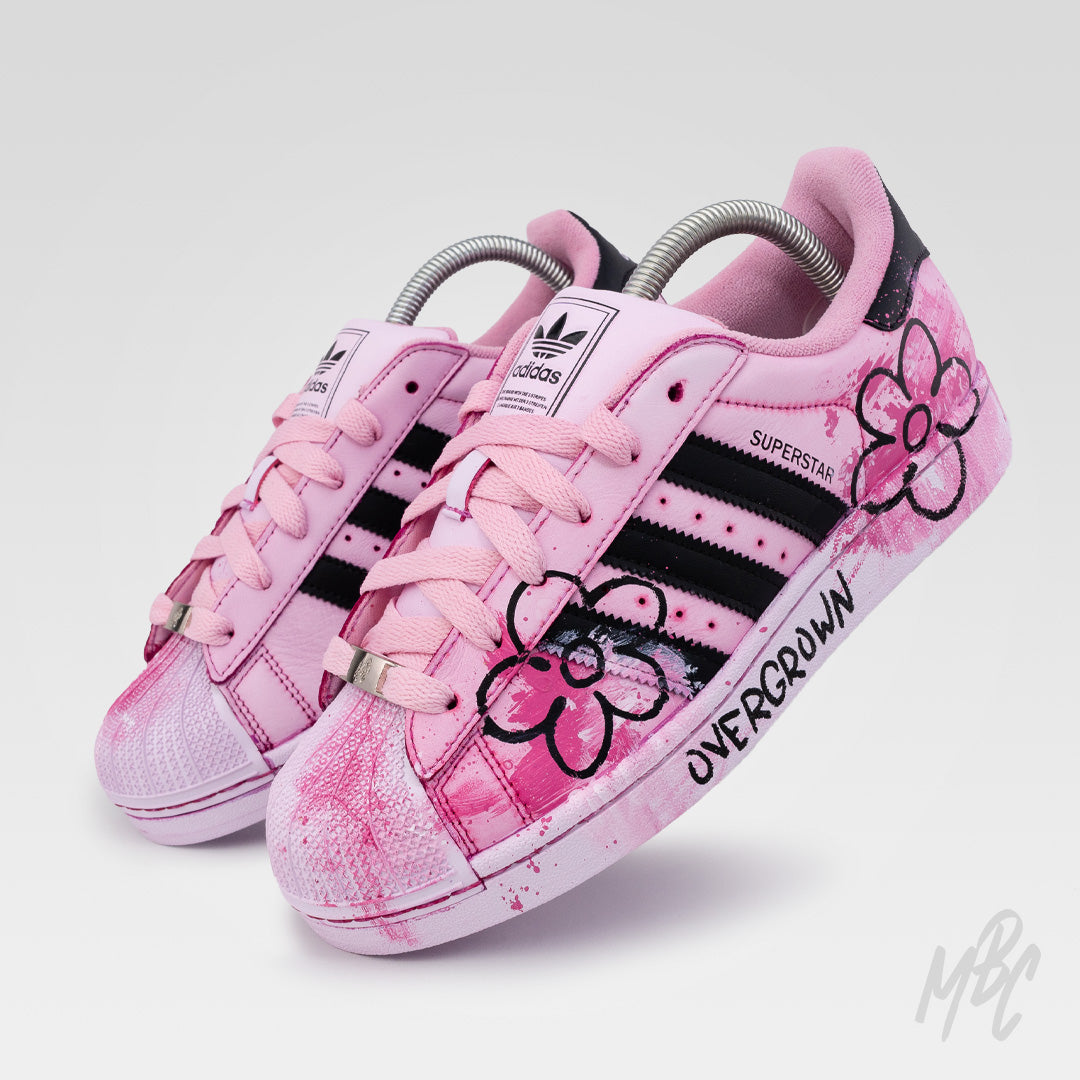Angled view of Pink Overgrown custom Adidas Superstar trainers with hand-painted floral graphics, pink paint texture and black Adidas stripes.
