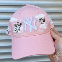 Pink Heaven New Era NY custom cap in pink with cherub illustrations and cloud detailing held outdoors
