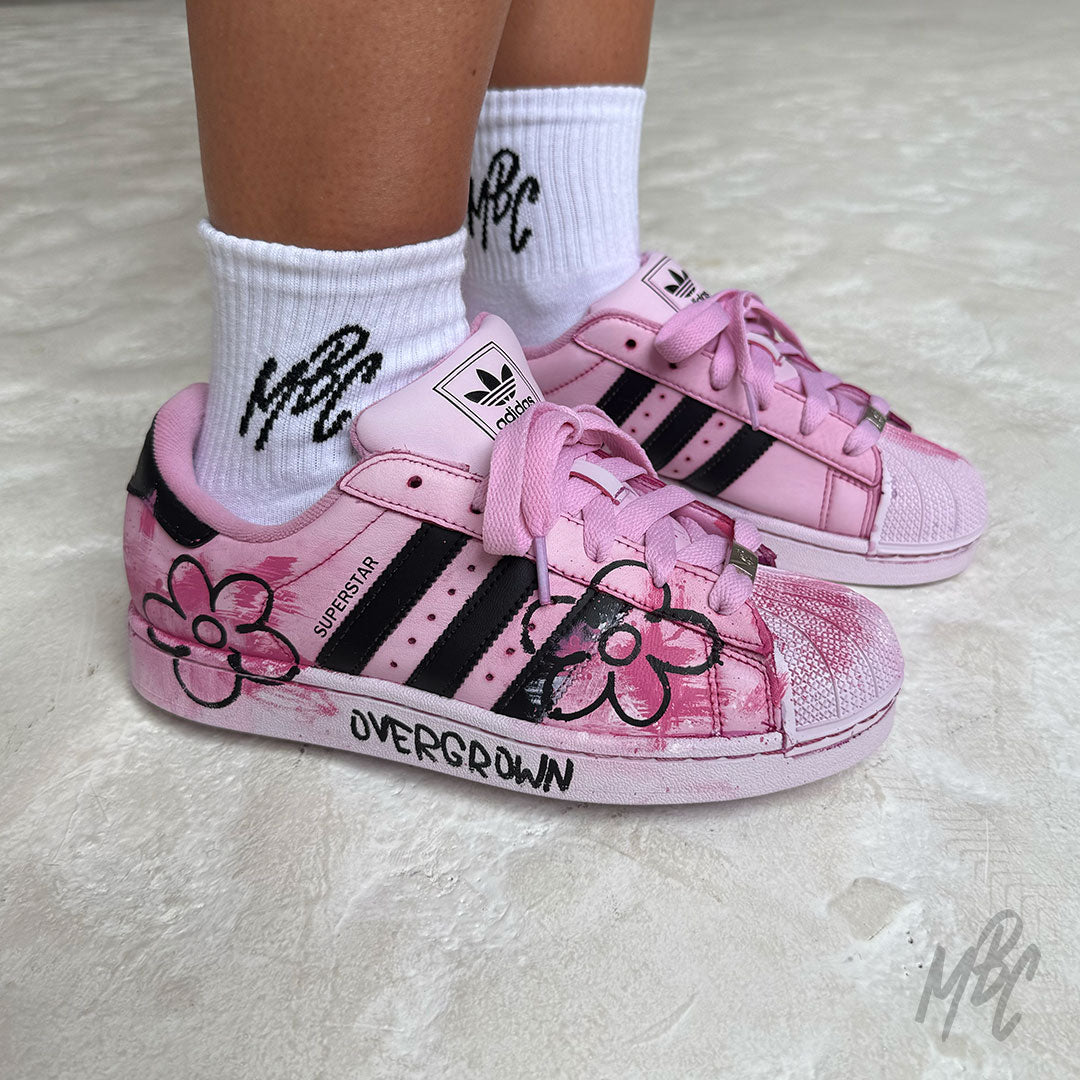 Pink Overgrown custom Adidas Superstar trainers worn on foot, featuring hand-painted pink brushstrokes, bold black floral artwork, painted midsoles and pink laces.