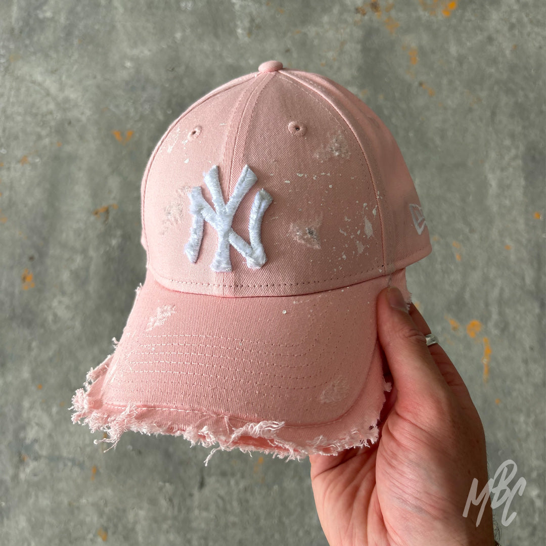 Pink Raw Edge custom New Era New York Yankees cap featuring a distressed frayed brim, frayed white embroidered NY logo, subtle white paint splatter detailing and a soft pink cotton finish, held in hand.
