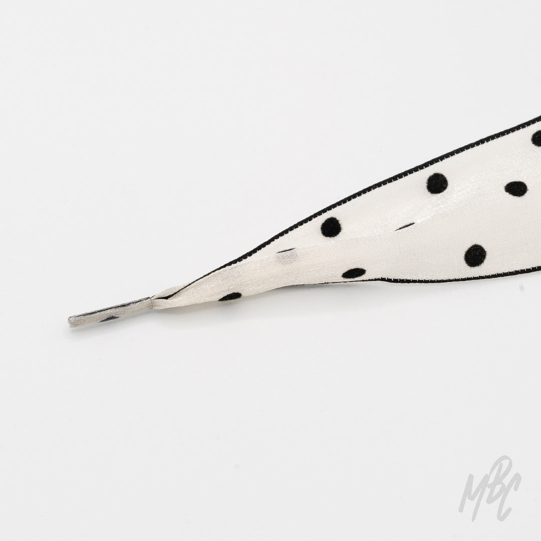 Close-up of a wide white fishtail ribbon shoelace with black polka dots, black edging, and a clear plastic aglet on a white background.