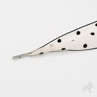Close-up of a wide white fishtail ribbon shoelace with black polka dots, black edging, and a clear plastic aglet on a white background.