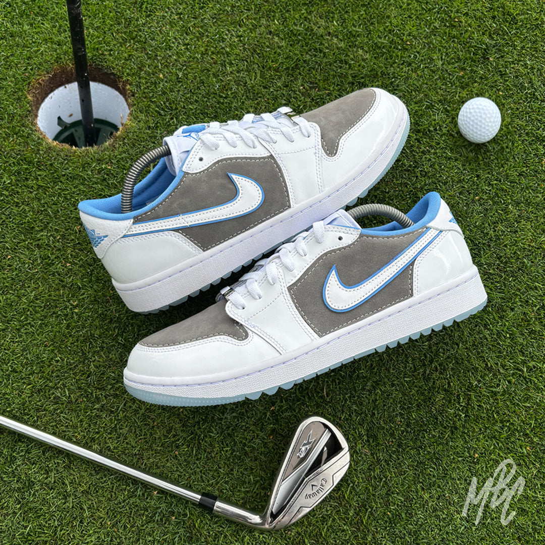 Pair of Slate Suede custom Jordan 1 Low Golf shoes with grey suede panels and light blue accents displayed on a putting green beside golf clubs and a golf ball.