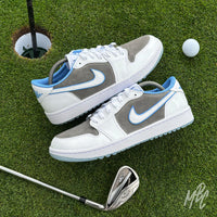 Pair of Slate Suede custom Jordan 1 Low Golf shoes with grey suede panels and light blue accents displayed on a putting green beside golf clubs and a golf ball.