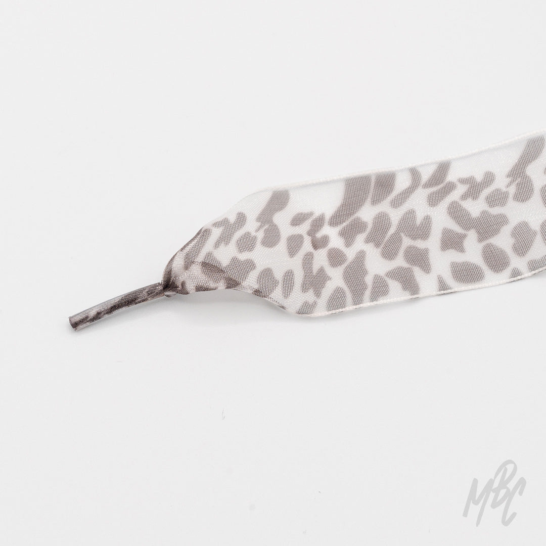 Close-up of a wide white ribbon shoelace with a gray snow leopard print and matching aglet on a white background.