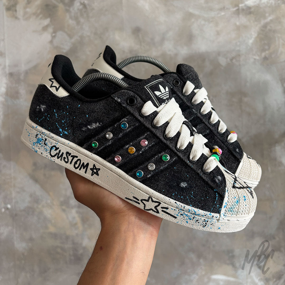Stardust Denim custom Adidas Superstar trainers featuring a black denim finish, colourful crystal detailing, hand-painted blue paint splatters, white rope laces and hand-drawn graphics.