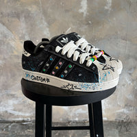 Stardust Denim custom Adidas Superstar displayed on a black stool, showcasing black denim panels, colourful crystal embellishments, hand-painted graphics and blue paint splatter detailing.